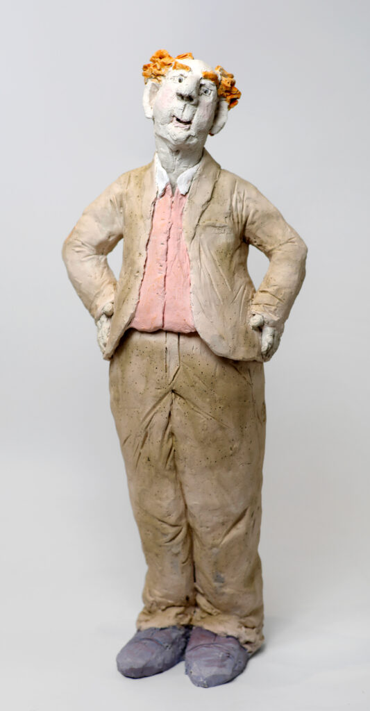 Sculpture figurative en terre cuite et engobes représentant un homme debout, mains sur les hanches, visage incliné, cheveux roux, chemise rose et haussures violettes