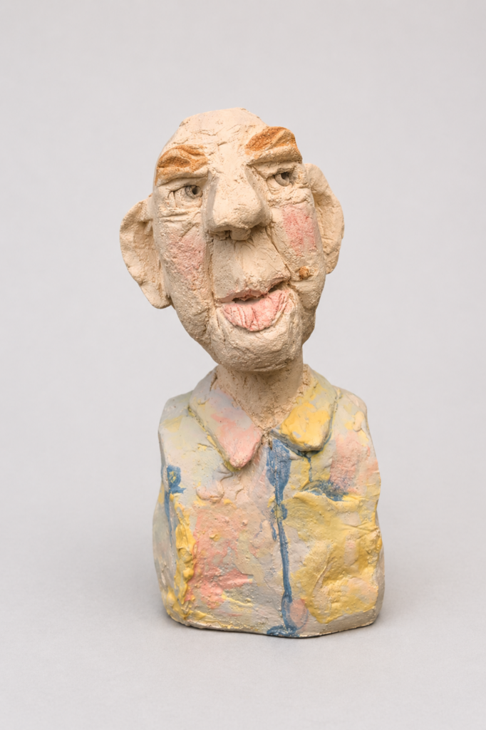 ^tête en grès chamotté et engobes d'un homme au visage allongé, bouche inférieure charnue, sourcils marqués, joues rosées et vêtement aux tons pastel jaune, rose et bleu